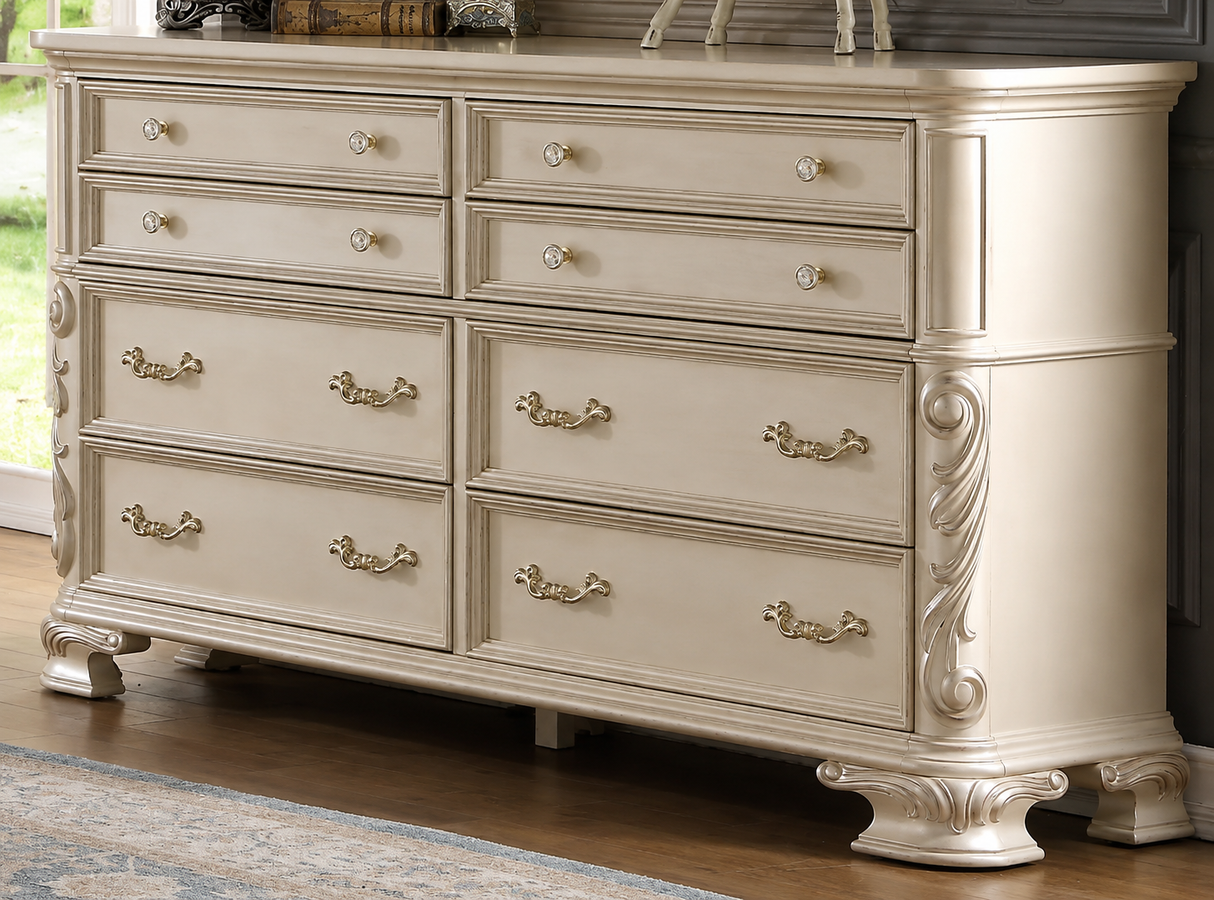 An eight-drawer dresser in a distressed cream or off-white finish with ornate silver metal handles and decorative carved side details, shown in a room setting with a wood floor and window.