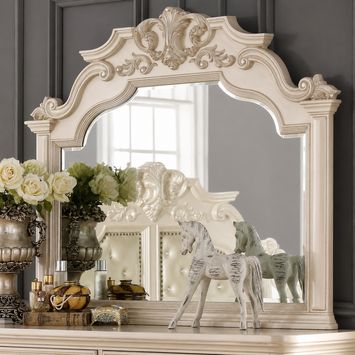 An ornate cream-colored wooden mirror with a highly decorative, classical scrollwork frame, displayed on a matching console table against a dark gray paneled wall. The scene is styled with vintage horse figurines, a floral arrangement, and decorative bottles.