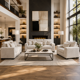 A staged living room featuring a light beige upholstered sofa, matching loveseat, and a rectangular coffee table on a large light rug, with a dark fireplace wall, built-in shelves, and tall windows in the background.