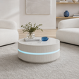 A modern round white coffee table with a ribbed lower section and a glowing blue LED light band, styled in a bright, minimalist living room with white armchairs and a light rug.