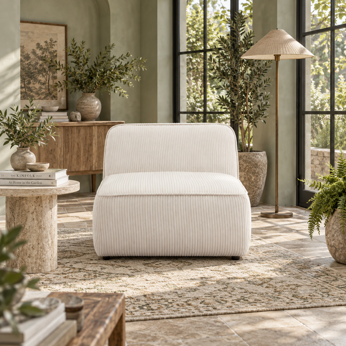 A modern upholstered accent chair in textured cream corduroy fabric, styled in a bright, sunlit room with olive green walls, a stone side table, and potted plants.