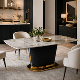 A modern dining table with a white marble top and a black quilted pedestal base accented by gold trim, surrounded by light beige upholstered chairs in a contemporary dining room setting.
