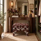 A styled vanity table setup in a warm room, featuring a dark wood writing desk with a matching upholstered stool, a gold dome lamp, and floral arrangements under a large mirror.