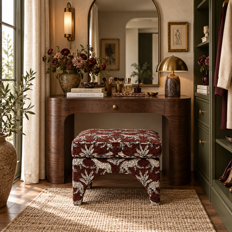A styled vanity table setup in a warm room, featuring a dark wood writing desk with a matching upholstered stool, a gold dome lamp, and floral arrangements under a large mirror.