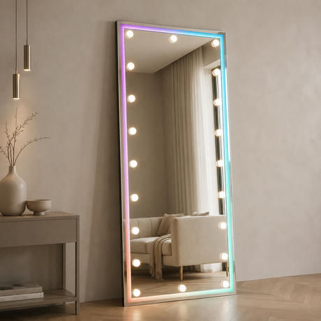A tall rectangular floor mirror with a slim silver frame and glowing multicolor LED light bulbs around the border, shown standing in a modern room with a side table and sofa reflected in the mirror.