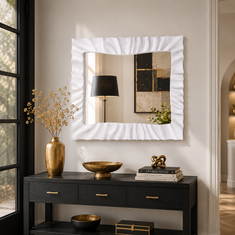 A white rectangular wall mirror with a ruffled, textured border hangs on a beige wall above a black console table decorated with gold bowls, a vase, and books. The mirror reflects a black lamp and framed art.