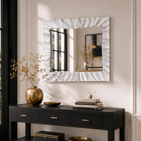 A square wall mirror with a textured, ruffled white frame hangs above a black console table decorated with a gold vase, books, and dried flowers in a styled interior setting.