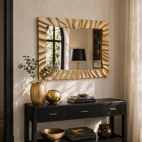 A large rectangular wall mirror with a textured, wavy gold frame hangs above a black console table decorated with gold vases, books, and bowls in a warm, styled interior setting.