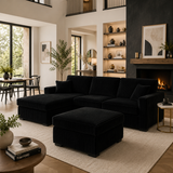 A modern black corduroy sectional sofa with a matching ottoman in a stylishly decorated living room with a fireplace, built-in shelves, and a dining area in the background.