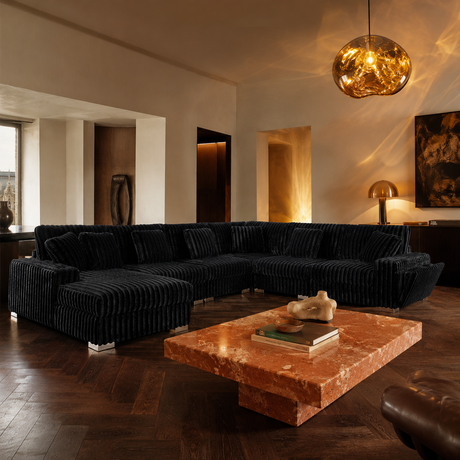 A luxury U-shaped sectional sofa upholstered in black ribbed corduroy fabric, styled in a modern, warm-toned living room with a terracotta marble coffee table and a glowing glass pendant light.