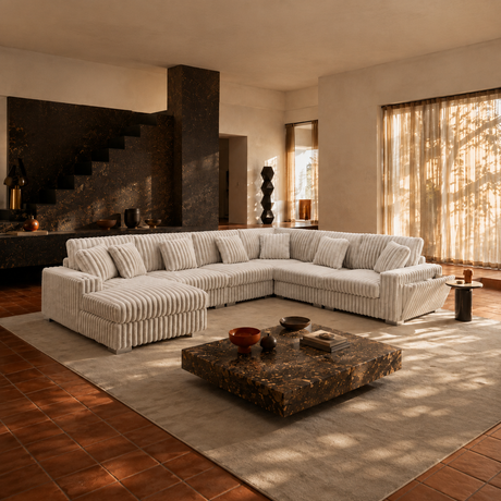 A large U-shaped sectional sofa upholstered in cream ribbed fabric, arranged in a modern living room with terracotta tile floors, a dark stone coffee table, and warm sunlight streaming through sheer curtains.