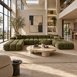 An olive green modular sectional sofa arranged in a U-shape in a modern, sunlit loft living room with a round stone coffee table and a large woven area rug.