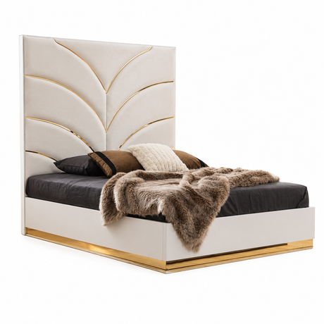 A modern platform bed with a tall tufted headboard in warm off-white leather, accented with gold leaf-like inlay lines and a gold metal base trim. The bed is styled with dark gray bedding, brown pillows, and a brown faux fur throw blanket, set against a plain white background.