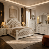 An ornate white carved bed frame with a tufted cream headboard and matching bedding, shown in a luxurious beige-toned bedroom with matching white furniture, lamps, and a large mirror.