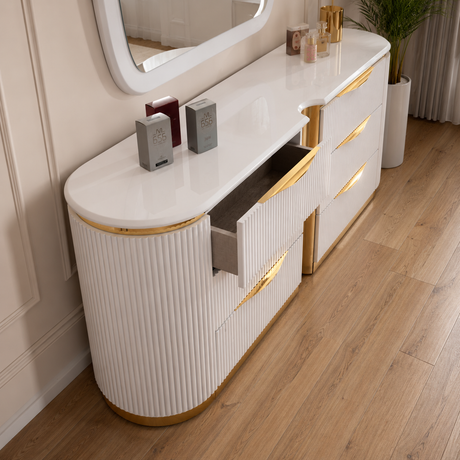 A modern white vanity or dresser with ribbed front panels, gold trim, and an open drawer, shown in a styled interior with wood flooring and wall decor.