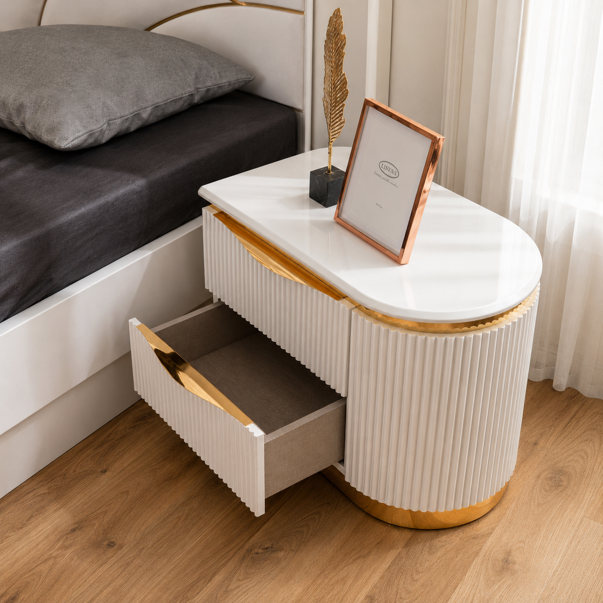 A modern oval bedside table with ribbed cream-white drawers, a glossy white top, and gold-toned metal trim, shown next to a bed in a bedroom. One drawer is open, and a decorative framed picture and feather stand sit on the tabletop.