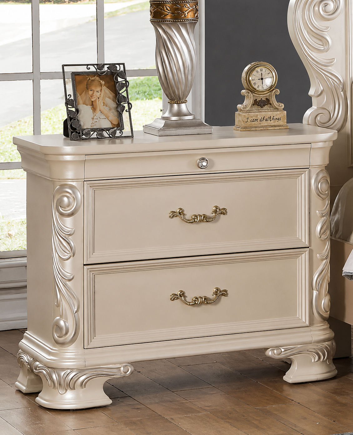 An ornate cream-colored two-drawer nightstand or end table with carved details and gold-toned handles, shown in a bedroom setting with decorative items on top.