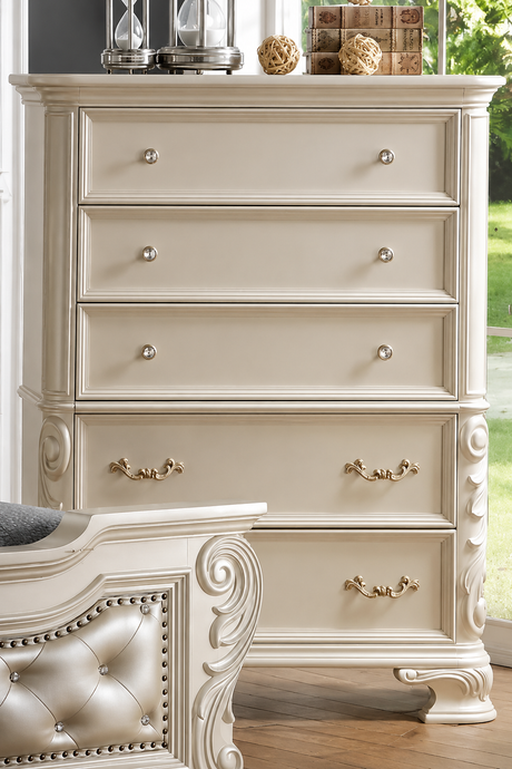 An ornate cream-colored wooden dresser with five drawers, featuring silver round knobs on the upper drawers and gold-toned bar handles on the lower drawers, set in a bedroom next to a matching tufted headboard.