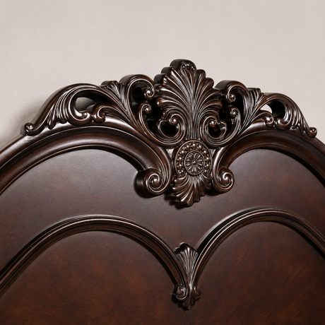 Close-up of an ornate dark brown wooden headboard with carved scrollwork and a central decorative crest, shown against a plain light beige wall.